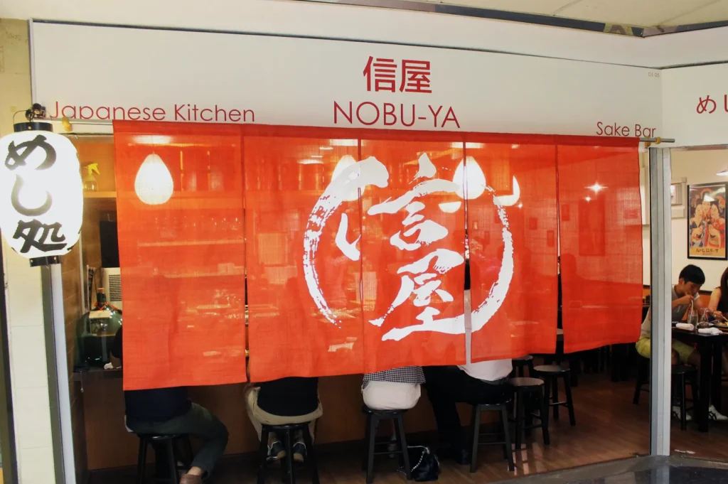 Japanese kitchen and sake bar with a red curtain featuring white kanji characters. Patrons sit inside and a lit lantern hangs outside, creating a cozy atmosphere.