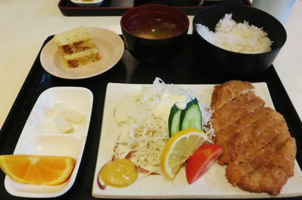 A Japanese meal set on a black tray includes breaded pork cutlet, shredded cabbage with cucumber and lemon, rice, miso soup, rolled omelet pieces, and orange slices.