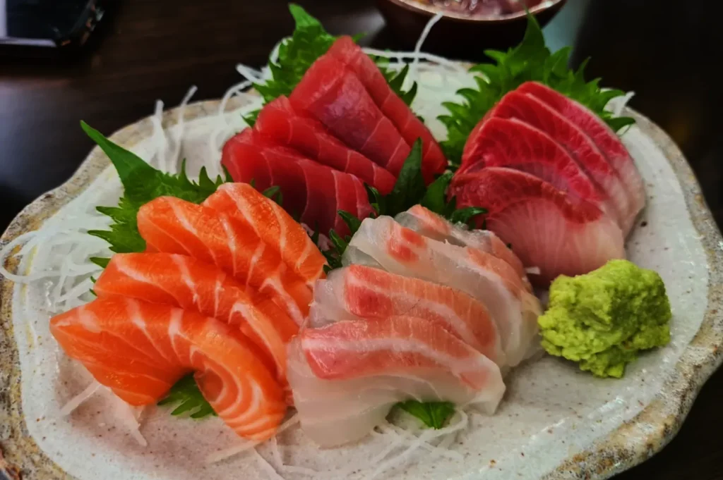 A plate of assorted sashimi featuring vibrant slices of salmon, tuna, and amberjack, garnished with green shiso leaves and a wasabi mound.