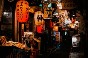 Dimly lit Japanese alley with glowing red and white paper lanterns and hanging decorations. Cozy, inviting atmosphere with small food stalls.