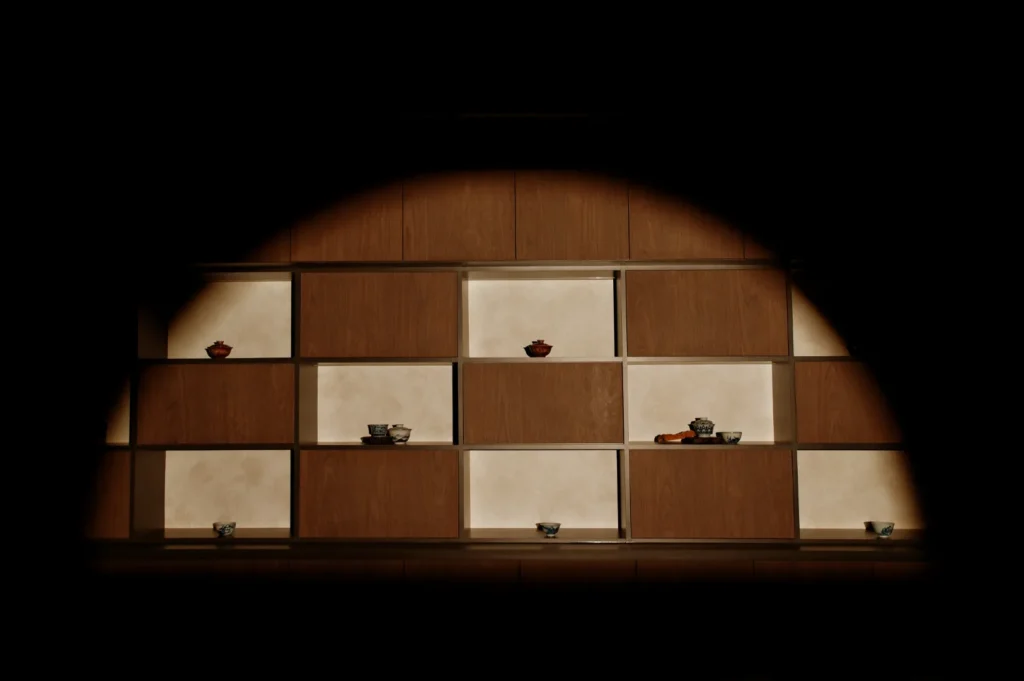 Wooden shelf with square compartments displaying small, assorted bowls and pots. Dim lighting creates a warm, intimate atmosphere.