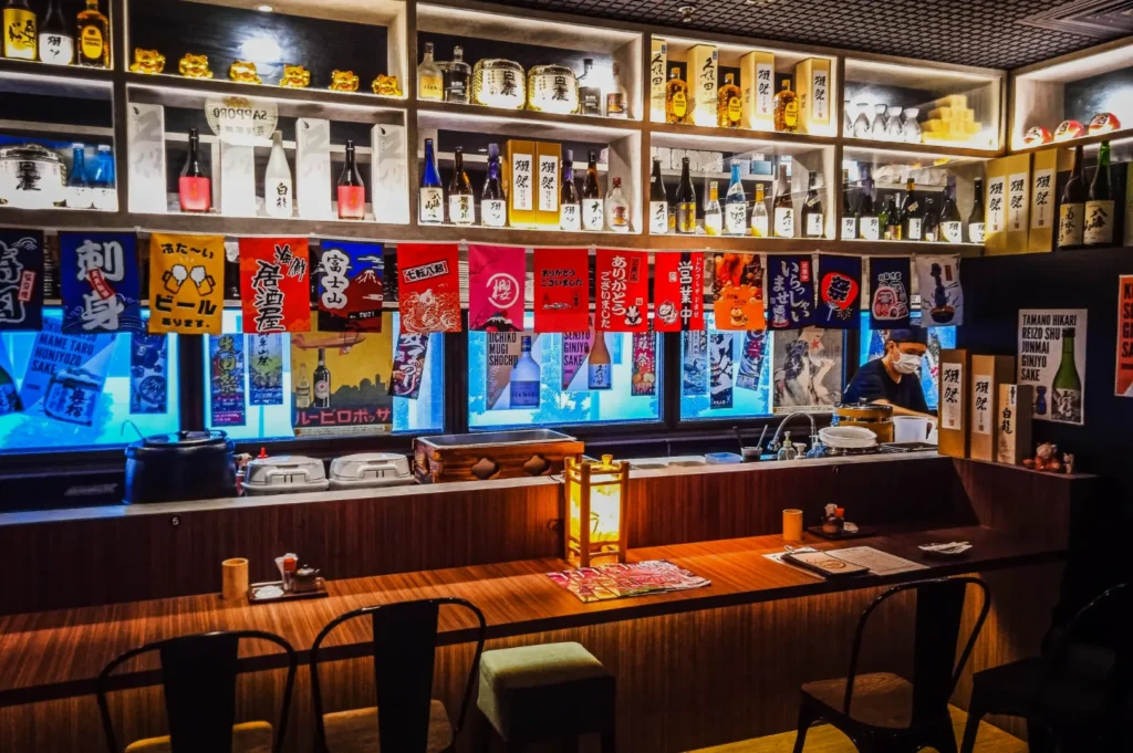 Cozy Japanese izakaya with shelves of sake bottles and colorful banners above a wooden counter. Ambient lighting, intimate atmosphere, staff working.