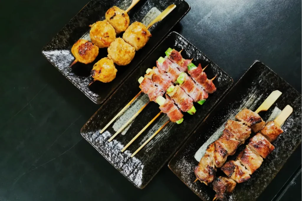 Three black plates with skewers of grilled meat. From left: chicken meatballs, bacon-wrapped vegetables, and beef cubes, creating a savory and appetizing display.