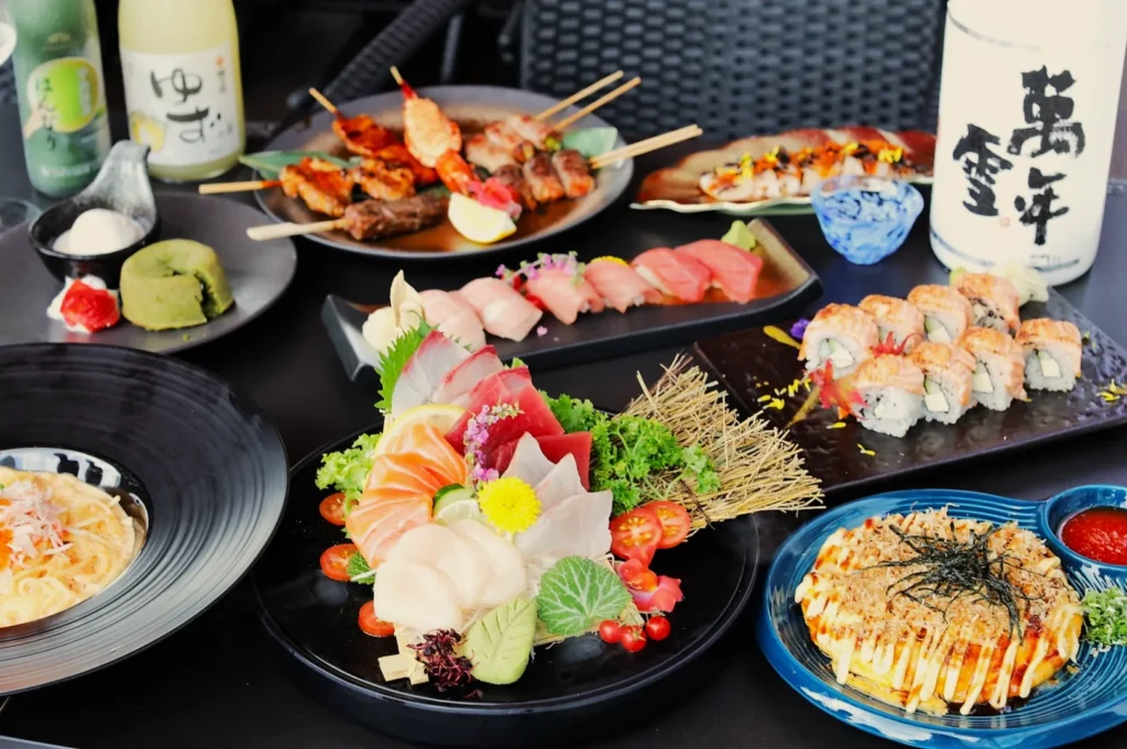 Assorted Japanese cuisine on a table, including sushi, sashimi, skewers, pasta, and okonomiyaki. Bottles of sake and vibrant garnishes.