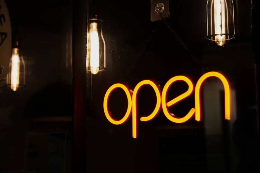 Bright neon "open" sign glows warmly in a dark setting, flanked by two vintage-style hanging bulbs, conveying a welcoming nighttime atmosphere.