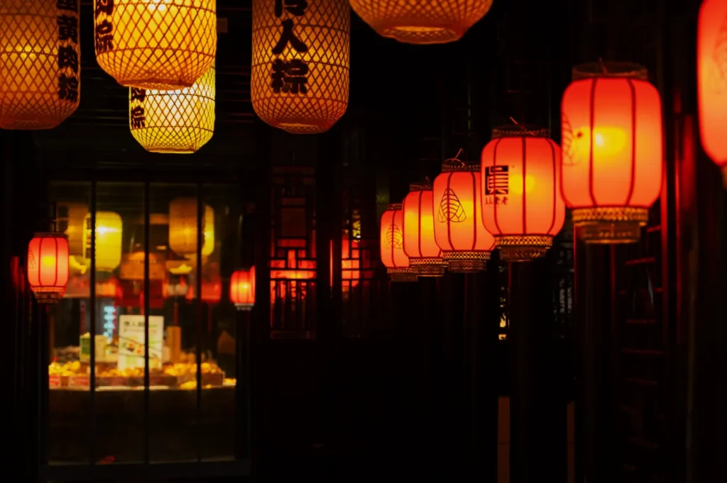 Row of glowing red and yellow Chinese lanterns line a dark corridor, casting a warm, inviting glow. The atmosphere is serene and traditional.