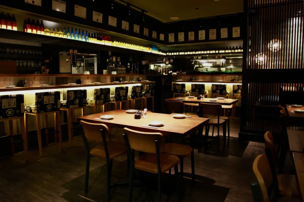 Cozy Japanese restaurant interior with dim lighting, wooden tables, and chairs. Bottles line the top shelf, and a partially open kitchen is visible in the back.