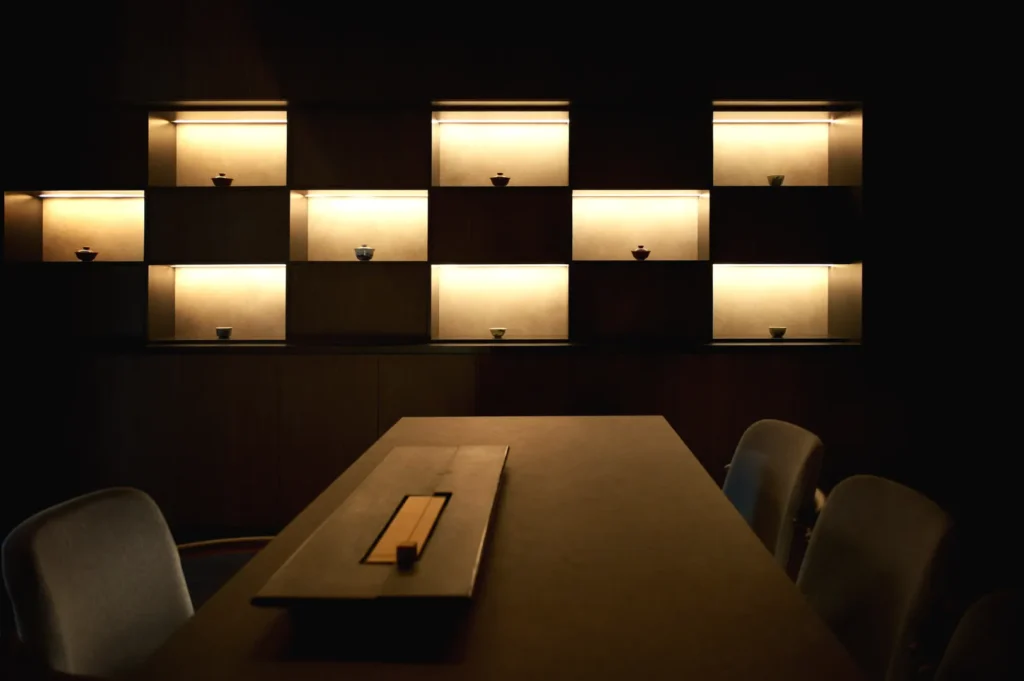 Dimly lit room with a wooden table and cushioned chairs. Illuminated shelves with small, decorative bowls create a cozy and minimalist ambiance.