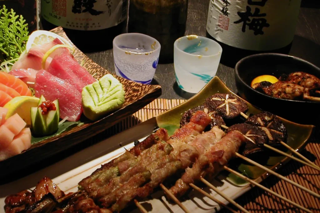 A beautifully arranged Japanese meal featuring assorted sashimi, yakitori skewers, and sake bottles. The setup conveys a warm, inviting dining experience.