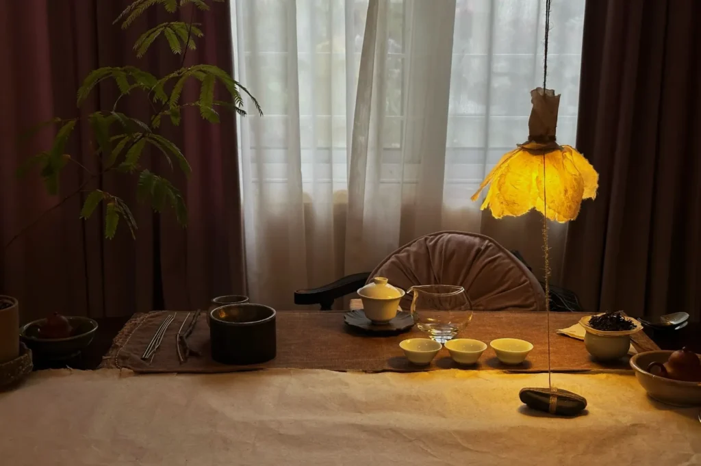 A cozy, dimly-lit dining table is set for tea with a teapot, cups, and bowls under a soft, glowing lamp. A green plant adds a natural touch.