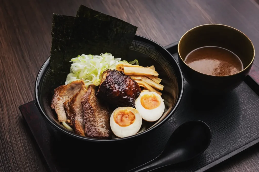 A bowl of ramen with sliced pork, halved soft-boiled eggs, bamboo shoots, shredded cabbage, and seaweed, served with soup on a wooden tray.