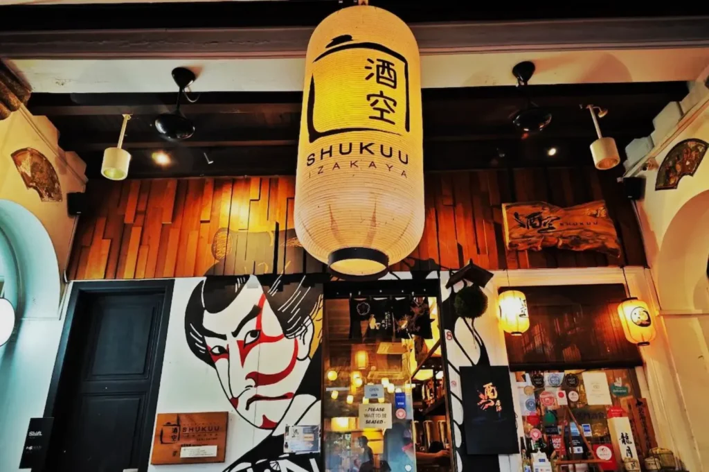 Large lantern with Japanese characters hangs above the entrance of Shukuu Izakaya. A bold mural of a face decorates the wooden facade. Cozy ambiance.