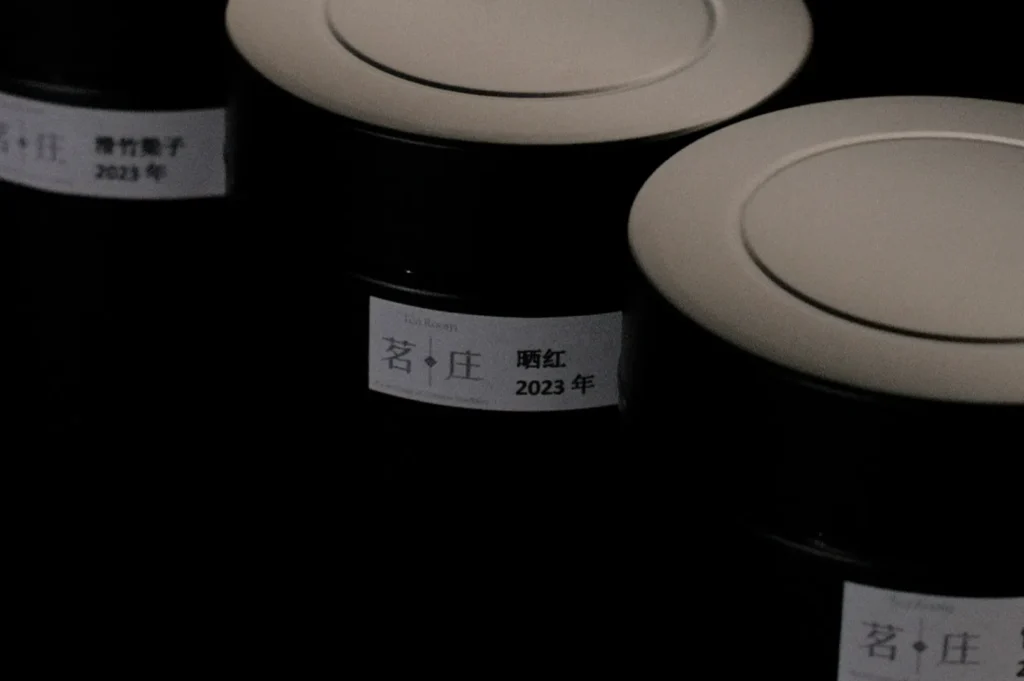 Dimly lit image of round tea canisters with beige lids, featuring labels with Chinese characters and "2023." The mood is calm and mysterious.