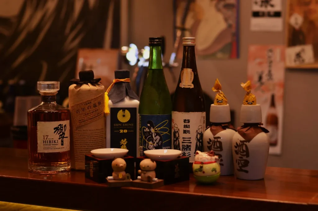 A collection of assorted liquor bottles, including whiskey and sake, on a wooden bar. The warm lighting creates a cozy, inviting atmosphere.