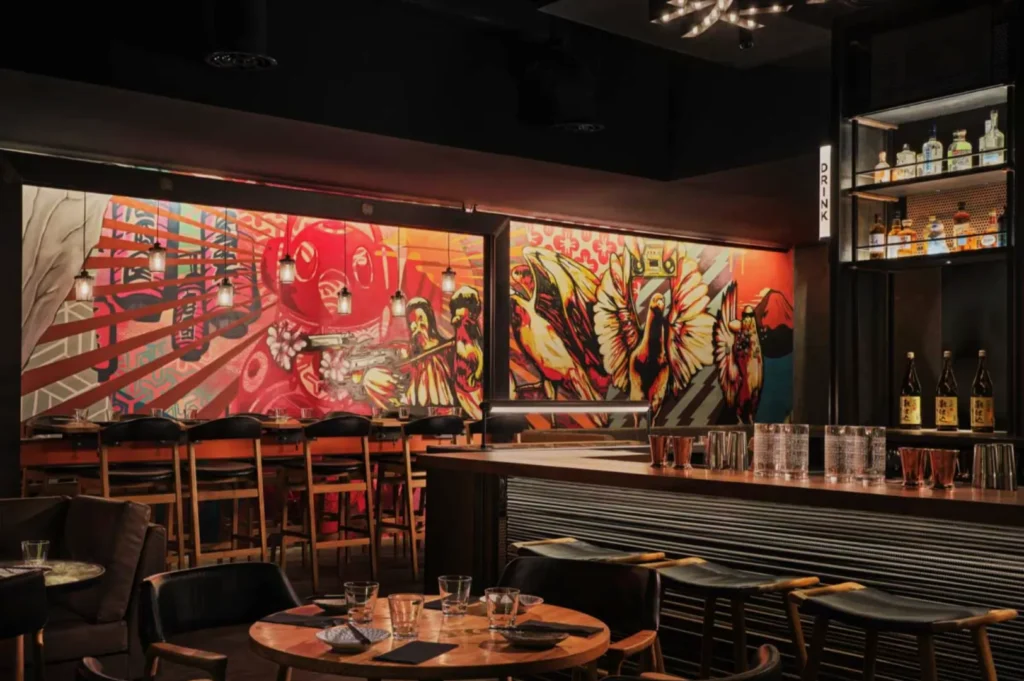 A dimly lit bar with colorful murals depicting abstract figures and birds. Wooden tables and stools create a warm, inviting atmosphere.