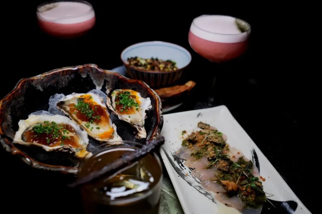 A gourmet spread featuring oysters topped with herbs and sauce, a dish of finely sliced fish, and vibrant pink cocktails, creating a luxurious mood.