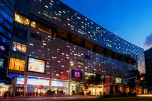 Modern shopping mall exterior at dusk, showcasing illuminated windows and signage. The vibrant blue sky and street scene add a lively urban atmosphere.