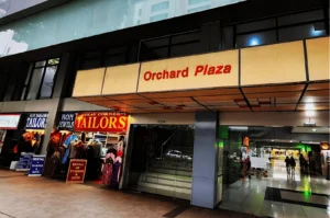 Storefront of Orchard Plaza with a sign. Tailor shops display colorful garments on the left, while indoor shopping is visible through glass doors.