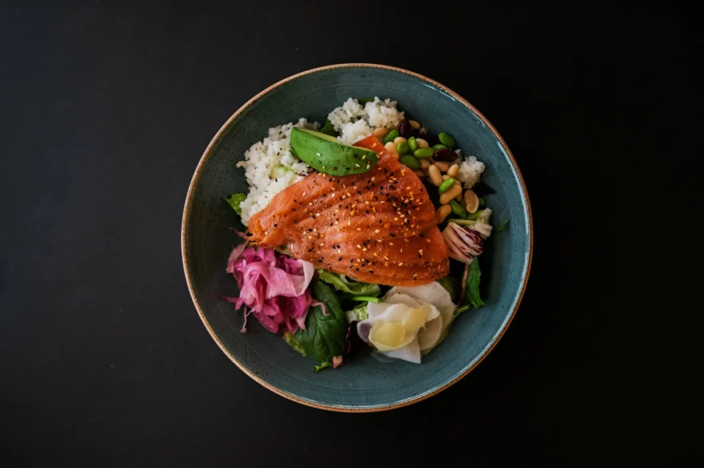 A bowl with vibrant, glazed salmon over rice, garnished with avocado, edamame, pickled vegetables, and greens, against a dark background.