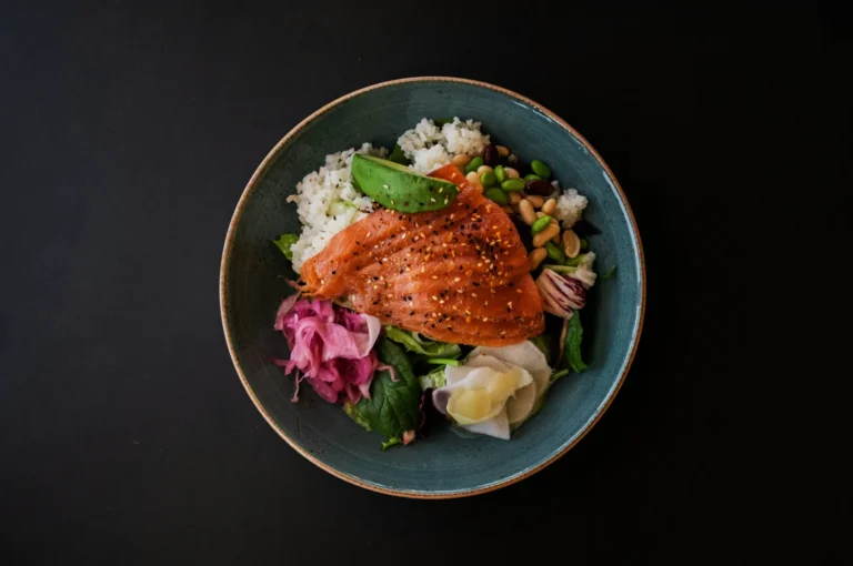 A bowl with vibrant, glazed salmon over rice, garnished with avocado, edamame, pickled vegetables, and greens, against a dark background.