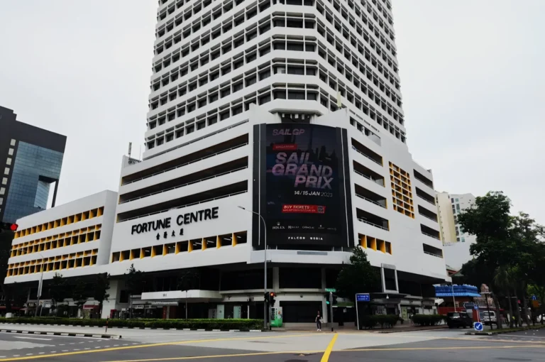 Tall white building with a "Fortune Centre" sign in front, showcasing a large "Sail Grand Prix" advertisement. Calm street scene with trees.