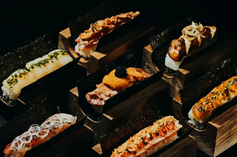Dark, moody close-up of assorted gourmet sushi hand rolls served in wooden holders, featuring toppings like uni, caviar, grilled fish, scallions, and crispy garnishes wrapped in seaweed.