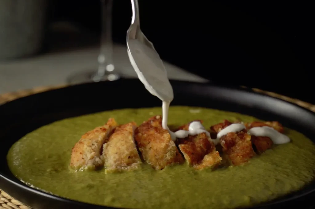 A low-angle shot captures a silver spoon drizzling a creamy white sauce over a sliced, golden-brown katsu cutlet. The crispy meat sits on a bed of smooth green curry sauce served inside a shallow black dish.