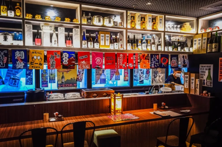 Cozy Japanese bar interior with wooden counter, colorful banners, and a variety of sake bottles on shelves. Warm lighting creates an inviting ambiance.