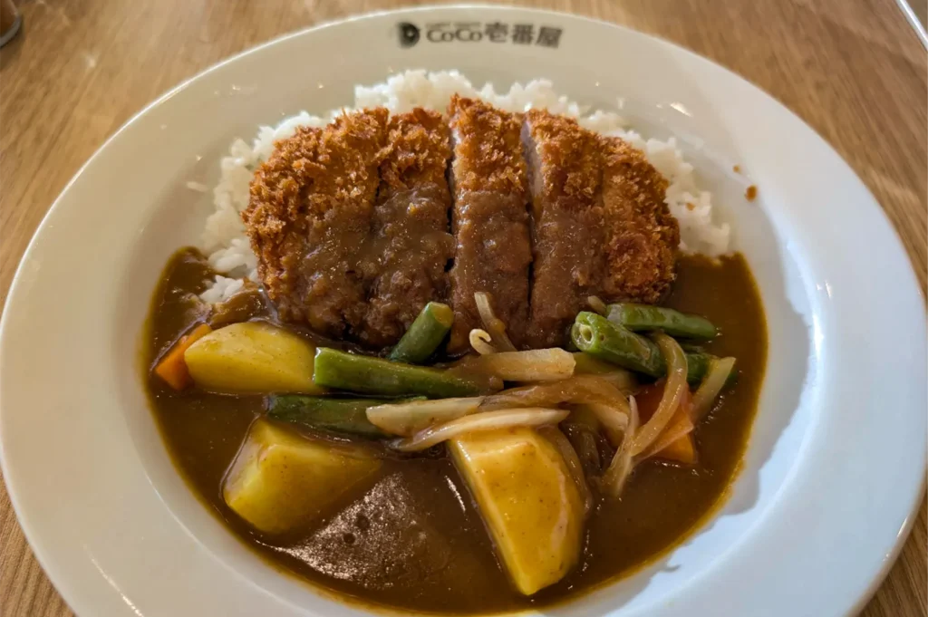 A high-angle shot captures a white bowl from CoCo Ichibanya filled with a bed of white rice and a rich brown curry sauce. The dish is topped with a sliced, crispy fried katsu cutlet alongside mixed vegetables including large chunks of potatoes, green beans, and onions.