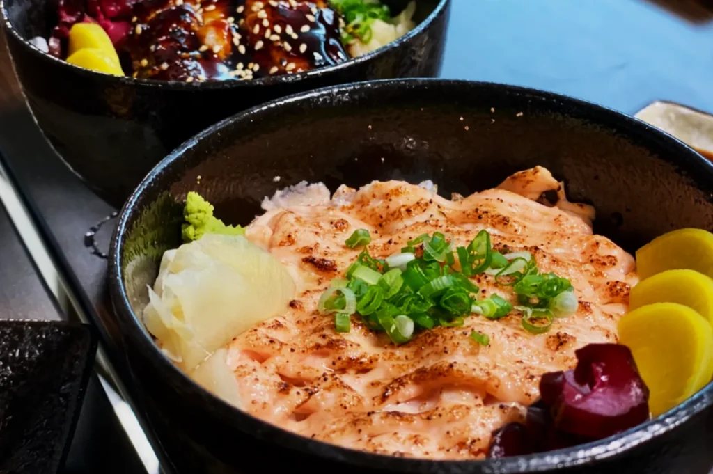Close-up of two Japanese rice bowls. The front bowl features creamy salmon topped with green onions, pickled ginger, and radish. The back bowl showcases glossy teriyaki chicken with sesame seeds. The bright colors and textures create an appetizing, vibrant scene.
