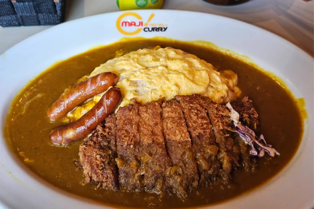 A high-angle shot captures a white Maji Curry plate filled with a large serving of Japanese curry sauce surrounding a mound of rice covered in a fluffy, swirled omelet. The dish is loaded with a sliced, crispy fried katsu cutlet in the foreground and two browned sausages resting on the side.