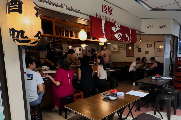 A cozy Japanese restaurant with wooden tables and stools, filled with people enjoying meals. Red lanterns and a welcoming atmosphere are present.