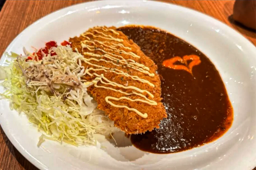 A high-angle shot captures a white plate filled with a large, crispy fried katsu cutlet drizzled with mayonnaise zigzags over a bed of white rice. The dish is accompanied by a dark, glossy curry sauce decorated with a small orange heart shape on one side, and a mountain of shredded cabbage salad on the other.