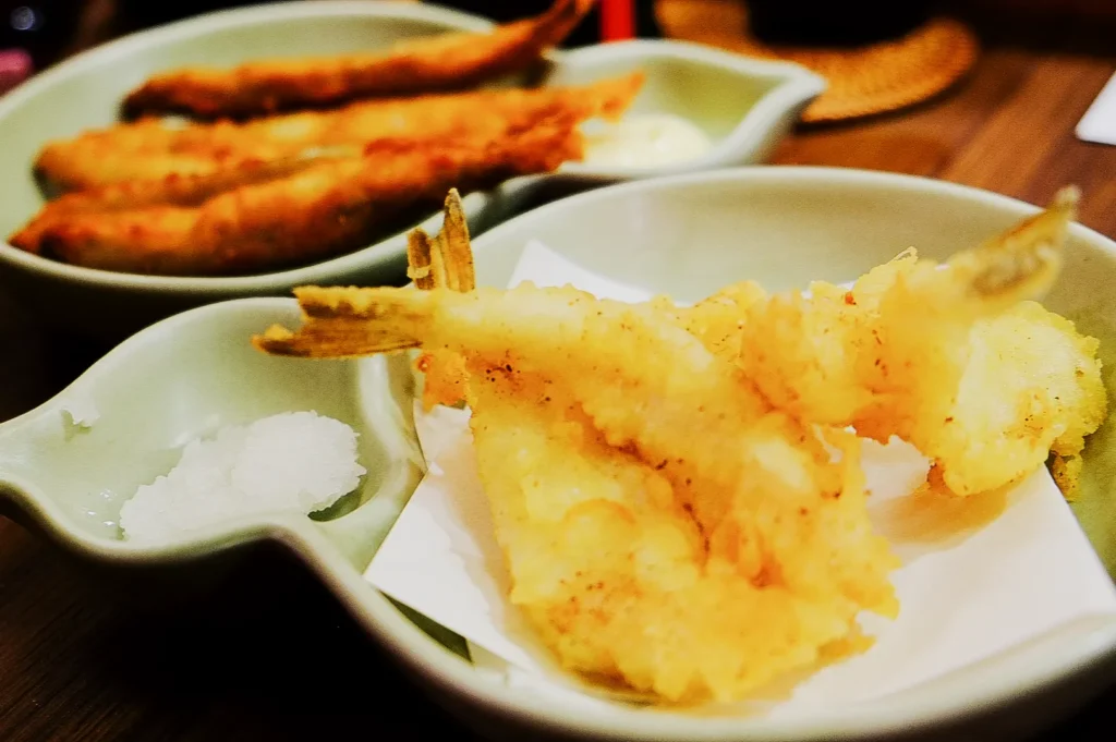 Golden tempura shrimp and fish are served on a green plate with a side of grated radish. The scene conveys a warm, appetizing tone. Keywords: tempura, shrimp, fish, Japanese cuisine.