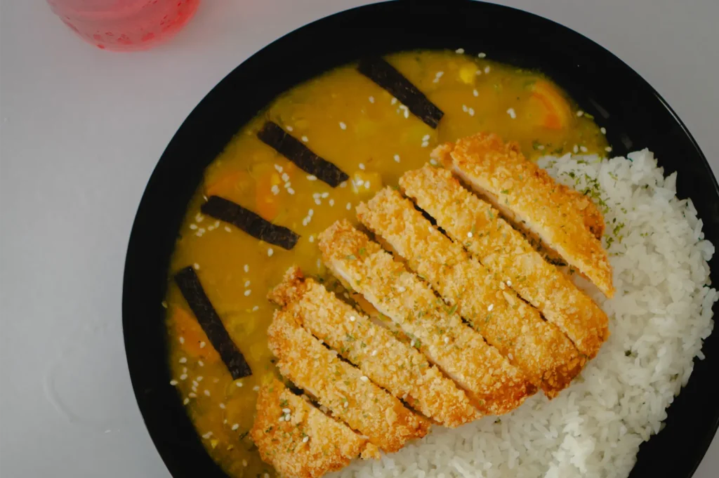 A top-down shot shows a black bowl filled with a side of white rice and a bright yellow curry sauce containing visible pieces of carrots. A sliced, golden-brown katsu cutlet rests across the middle, garnished with white sesame seeds and flanked by small strips of nori seaweed arranged like clock markers in the sauce.