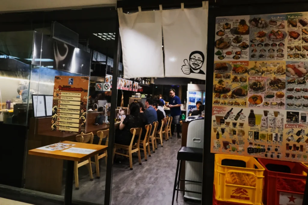 Cozy Japanese restaurant interior with wooden chairs, patrons dining, and a waiter in a blue shirt. Menu posters display colorful food images by the entrance.