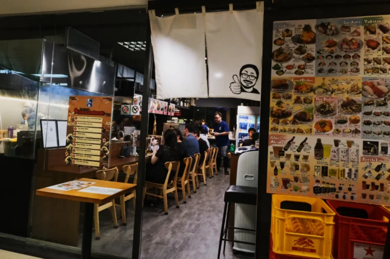 Cozy Japanese restaurant interior with wooden chairs, patrons dining, and a waiter in a blue shirt. Menu posters display colorful food images by the entrance.