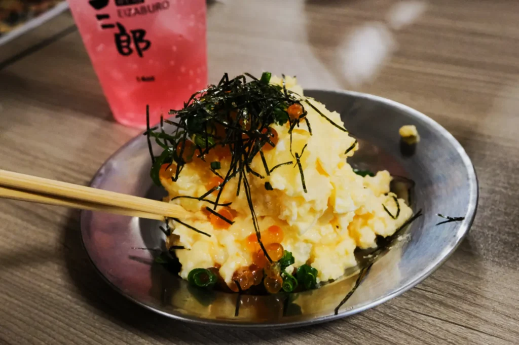 A metal plate with creamy scrambled eggs topped with seaweed, green onions, and salmon roe. Chopsticks rest on the plate. A pink drink is nearby.