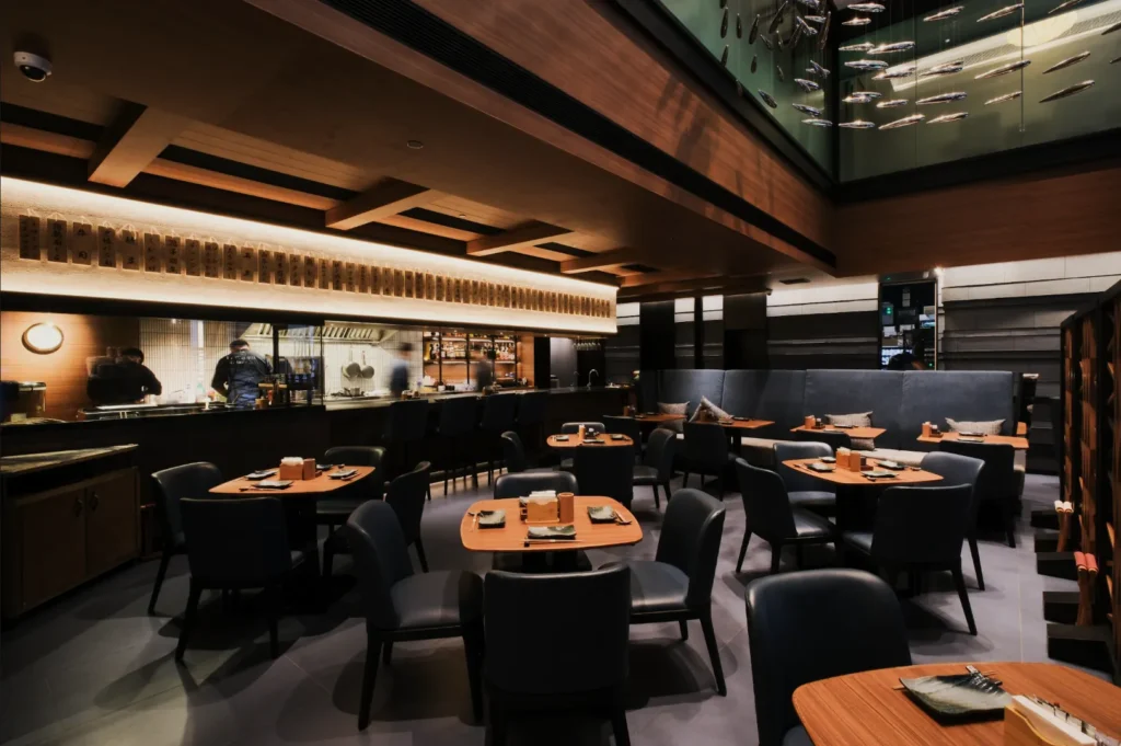 Modern restaurant with wooden tables and dark chairs, softly lit by overhead lights. Open kitchen and bar at back, creating a warm, inviting atmosphere.