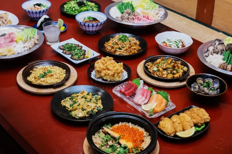 A variety of Japanese dishes are elegantly arranged on a wooden table. Plates of sushi, tempura, sizzling noodles, and fresh vegetables create a vibrant, inviting spread.