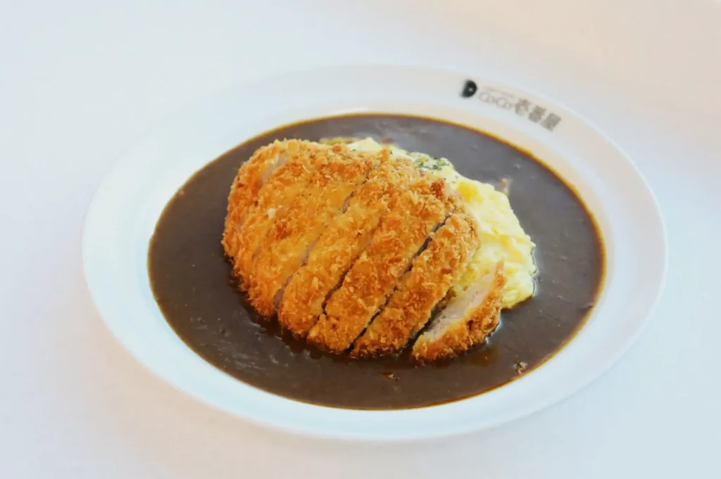 A white plate with crispy, golden-brown katsu slices atop creamy omurice covered in rich, dark curry sauce, creating a savory, comforting meal.
