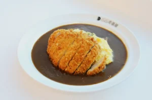 A white plate with crispy, golden-brown katsu slices atop creamy omurice covered in rich, dark curry sauce, creating a savory, comforting meal.