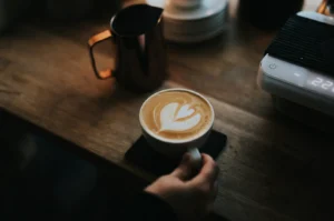 A hand holds a cup of latte with heart-shaped foam art on a wooden table, beside a metal milk frothing jug and a sleek espresso machine. Cozy ambiance.