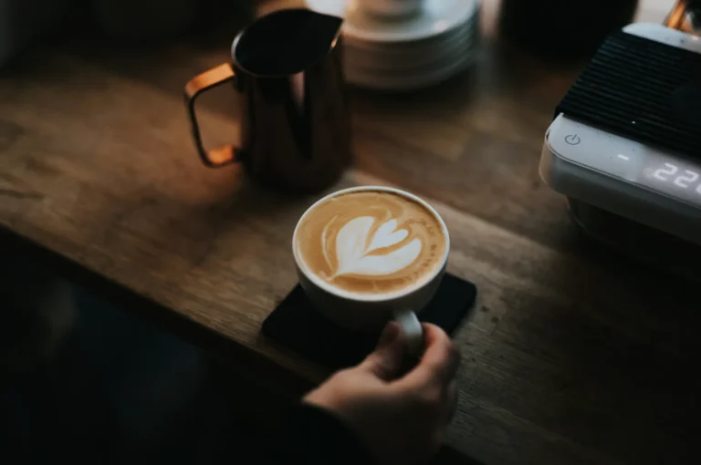 A hand holds a cup of latte with heart-shaped foam art on a wooden table, beside a metal milk frothing jug and a sleek espresso machine. Cozy ambiance.
