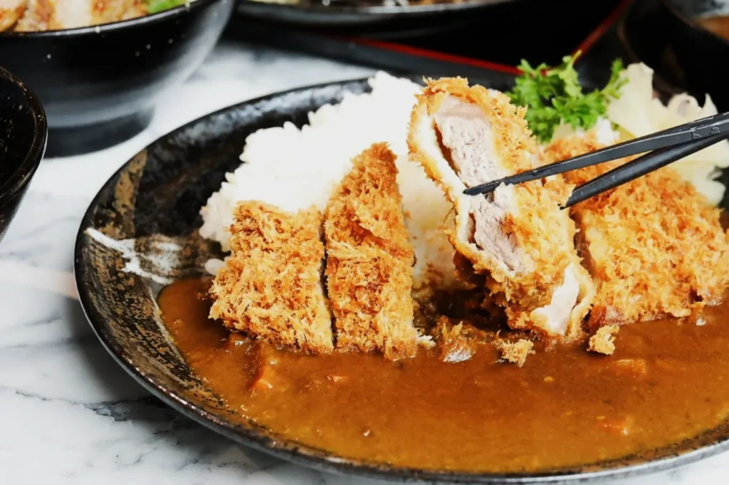 A plate of Japanese curry with rice and crispy breaded pork cutlet, garnished with parsley. Chopsticks hold a piece, emphasizing the dish’s texture.