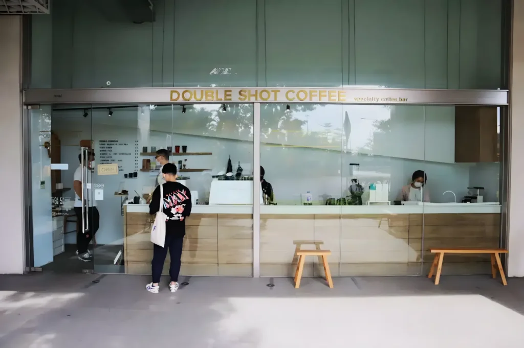 Exterior of "Double Shot Coffee" shop with glass facade. Two people inside, one outside. Minimalist design, wooden accents, calm and inviting atmosphere.