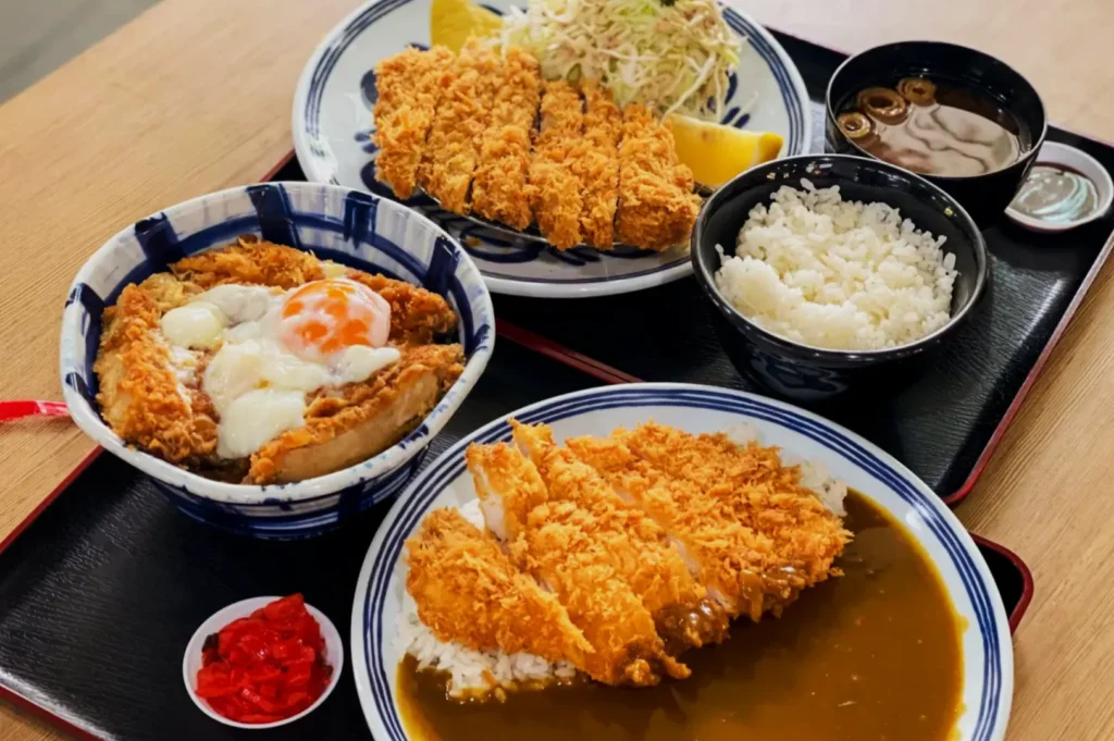 A delicious Japanese meal featuring katsu dishes: a bowl with egg-topped katsu, another with curry katsu, and a katsu plate with cabbage, served with rice and soup.