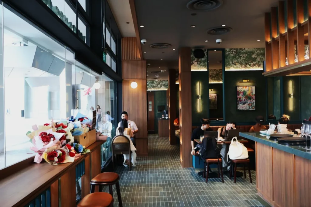 Stylish cafe interior with modern decor, teal and wood accents. Patrons sit at tables, a counter with flowers on the left. Bright, cozy atmosphere.