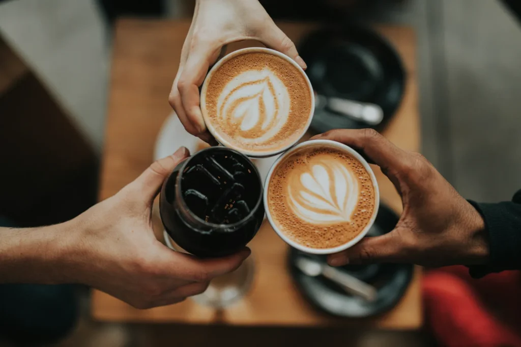 Three hands holding drinks in a toasting gesture: two lattes with heart-shaped latte art and one iced coffee. The scene conveys warmth and camaraderie.
