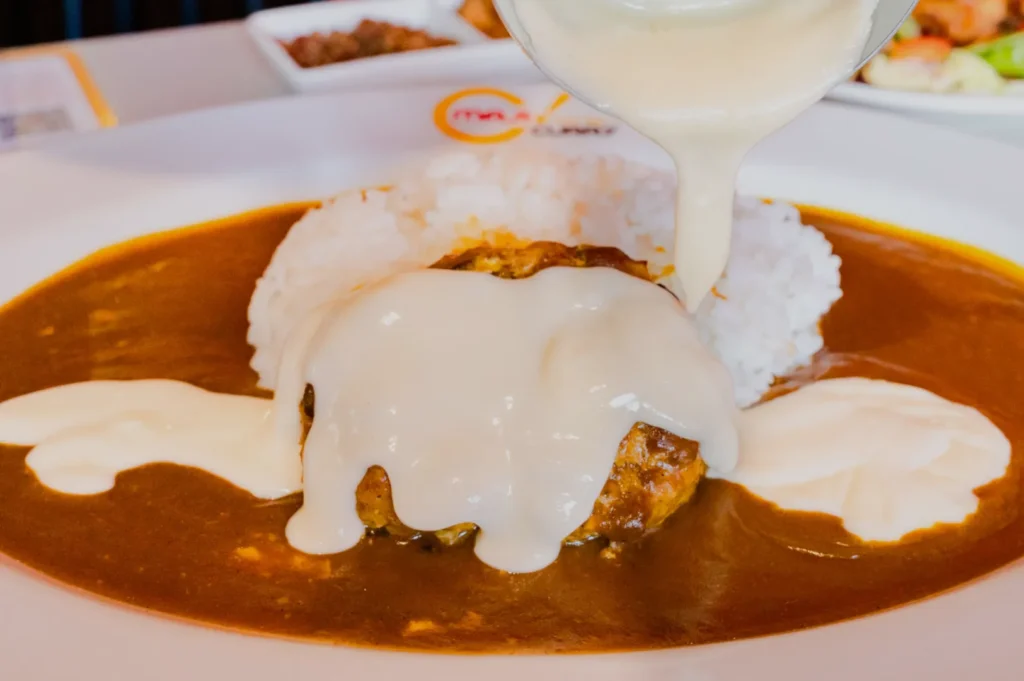 A plate of curry rice is topped with a meat patty, over which creamy white sauce is being poured, creating a rich and appetizing appearance.